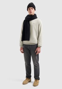 Beige sweatshirt, zwarte ribgebreide sjaal, zwarte jeans, beige laarzen. Model staat tegen een lichtgrijze achtergrond, met een neutrale uitdrukking.