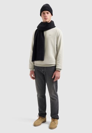 Beige sweatshirt, zwarte ribgebreide sjaal, zwarte jeans, beige laarzen. Model staat tegen een lichtgrijze achtergrond, met een neutrale uitdrukking.