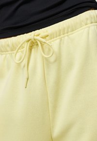 Gros plan sur un pantalon de survêtement jaune pâle avec une taille élastique et un cordon noué, porté avec un haut noir.
