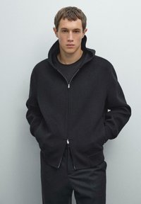 Jongeman met kort haar, gekleed in een zwarte zip-up hoodie over een zwart T-shirt, met zijn handen in de zakken, staande tegen een effen grijze achtergrond.