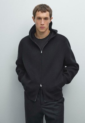 Giovane uomo con capelli corti che indossa una felpa nera con zip sopra una maglietta nera, con le mani nelle tasche, in piedi contro uno sfondo grigio uniforme.