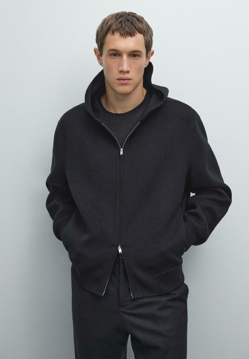 Jongeman met kort haar, gekleed in een zwarte zip-up hoodie over een zwart T-shirt, met zijn handen in de zakken, staande tegen een effen grijze achtergrond.