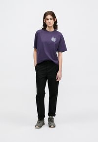 Fioletowa bawełniana koszulka z małą grafiką, w zestawieniu z czarnymi spodniami i szarym obuwiem o teksturowej powierzchni i kontrastowej podeszwie.