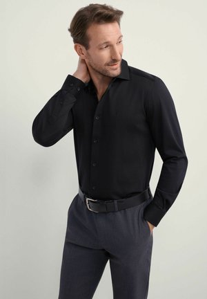Czarna koszula z długim rękawem, ze strukturalnym kołnierzem, gładka w dotyku. Sparowana z szarymi spodniami i czarnym paskiem, podkreślająca formalny styl.