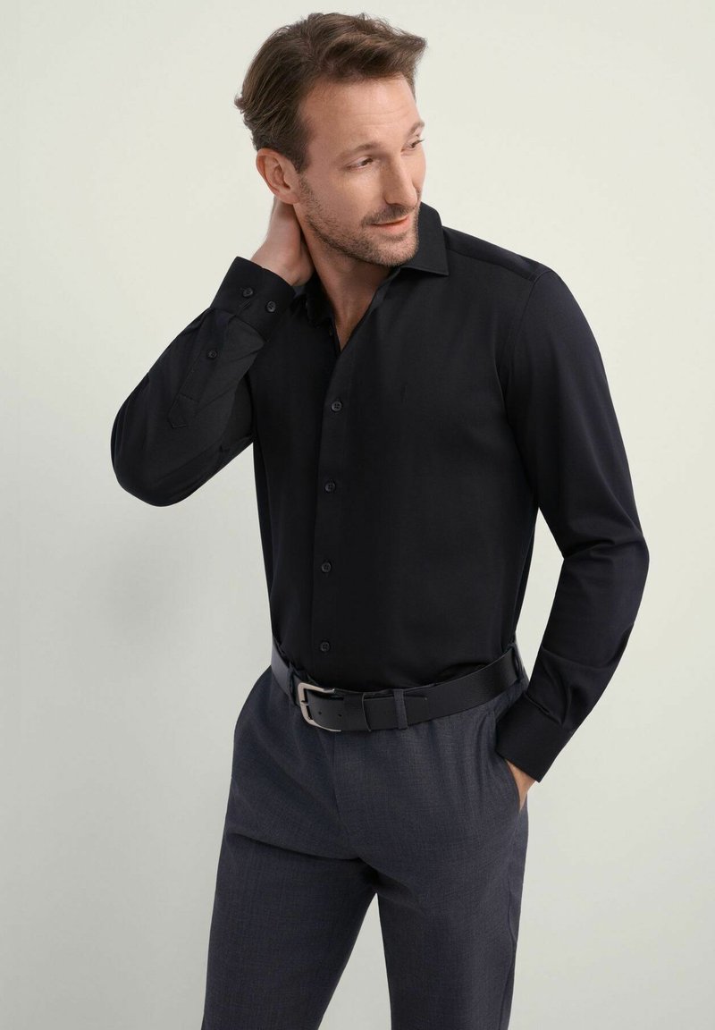 Czarna koszula z długim rękawem, ze strukturalnym kołnierzem, gładka w dotyku. Sparowana z szarymi spodniami i czarnym paskiem, podkreślająca formalny styl.