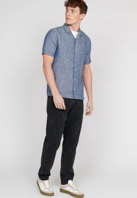 Camicia blu in chambray a maniche corte con colletto, dal tessuto texture, abbinata a pantaloni neri e sneakers bianche con dettagli verdi.