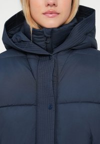 Jachetă puffer de marină cu guler înalt, detalii cusute, fermoar frontal și închidere cu butoane. Țesătura are un finisaj neted și texturat.