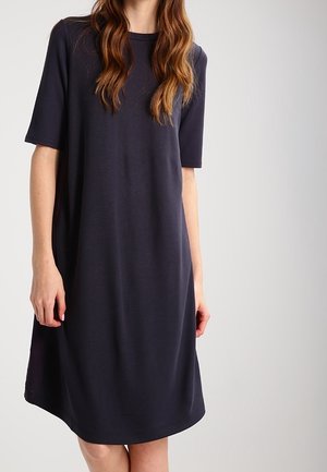 Woman wearing a loose-fitting, knee-length navy blue dress with short sleeves, standing against a plain light grey background.