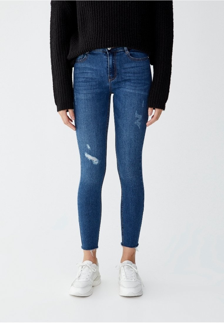 Personne portant un jean skinny bleu avec de légers détails usés, un pull noir à manches longues et des baskets blanches, debout devant un fond uni.