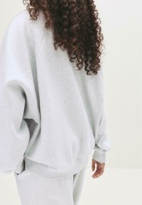 Person mit langen lockigen Haaren, die einen hellgrauen übergroßen Sweatshirt und passende Jogginghosen trägt, vor einem schlichten hellen Hintergrund stehend.
