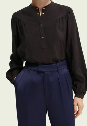 Sort bluse med høj halsudskæring, guldknapdetaljer og puffede ærmer, kombineret med navyblå plisserede bukser. Glat stoftekstur.