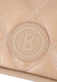 Beżowa skórzana torebka pikowana, z okrągłą naszywką z logo z literą "B" na środku. Podkreślona kontrastującymi detalami przeszycia.