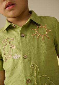 Chemise verte à manches courtes avec un tissu texturé. Présente des motifs brodés de soleil et de dinosaure en orange et blanc. Boutons en bois.