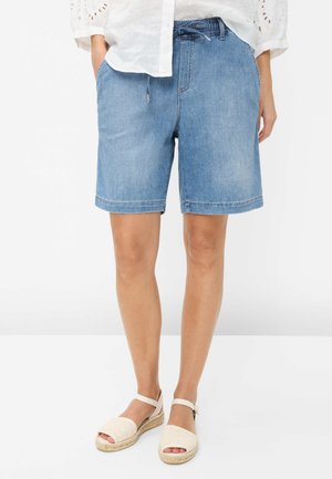 Kobieta ubrana w jasnoniebieskie dżinsowe szorty z elastycznym paskiem i białe sandały espadryle, stojąca na gładkim tle.