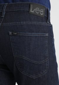 Mörkblå denimjeans med en synlig märke i midjan. Har två bakfickor och kontrasterande söm-detaljer.