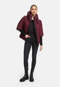 Burgunderfarbener Puffer-Jacke mit Kapuze, verfügt über einen Reißverschluss, kurze Ärmel und ein gestepptes Design, kombiniert mit schwarzen Skinny-Jeans und schwarzen Stiefeln.