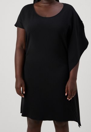 Kvinde iført en sort asymmetrisk kjole med rund halsudskæring og et kort ærme, stående foran en ensfarvet baggrund.