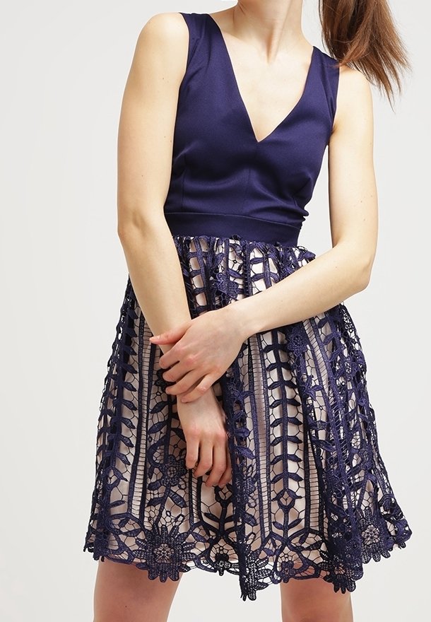 Žena v modrém bezrukávovém topu a šatech s květinovou krajkovou sukní, stojí s přehnutýma rukama před jednobarevným pozadím.