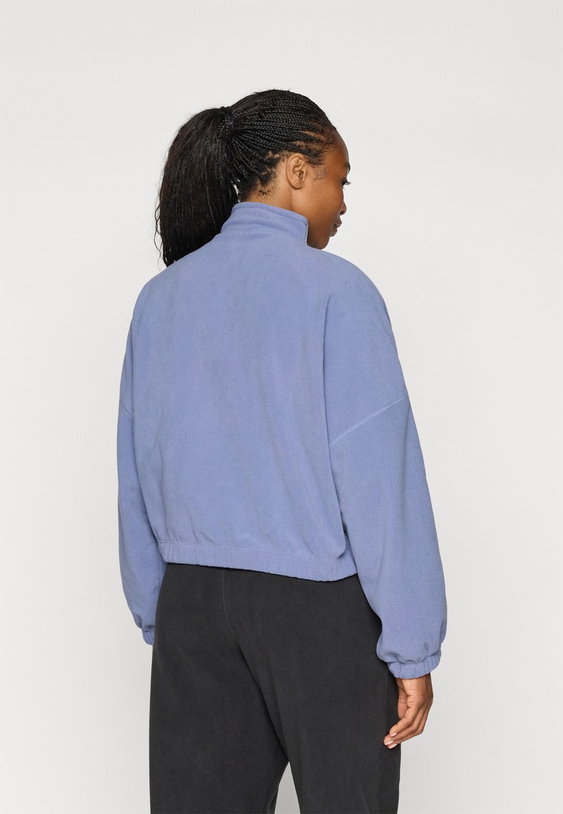 Fleecejacke mit hohem Kragen, in Hellblau, mit überschnittenen Schultern und elastischen Bündchen, getragen über schwarzen Hosen.