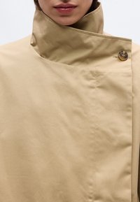 Εγγύς πλάνο ενός ατόμου που φοράει μπεζ παλτό με ψηλό γιακά, σφραγισμένο με ένα καφέ κουμπί κοντά στον λαιμό.