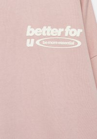 Tissu rose clair avec texte blanc indiquant "better for u" et "be more essential" à l'intérieur d'une forme ovale dans la partie supérieure.