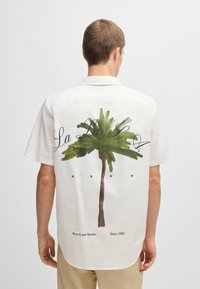 Korteblauwe witte shirt van lichtgewicht stof, met een groene palmboomafbeelding en zwarte tekst op de achterkant.