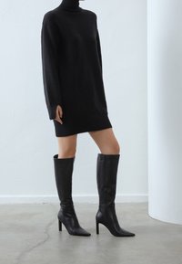 Robe pull noire à col roulé avec un ourlet côtelé, associée à des bottes cuissardes en cuir noir à talons aiguilles, dotées d'une fermeture éclair sur le côté.