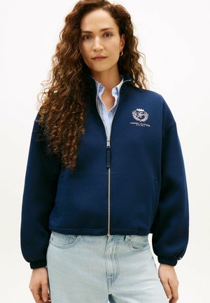 Frau mit langen, lockigen Haaren, die eine marineblaue Jacke mit Reißverschluss und einem Tommy Hilfiger-Logo sowie hellblaue Jeans trägt.