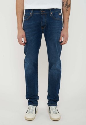 Man in blauwe rechte jeans, witte sneakers en een wit overhemd, staand met ontspannen armen langs het lichaam tegen een effen achtergrond.