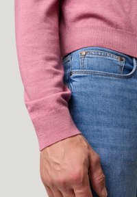 Rosa Pullover mit gerippten Bündchen, getragen über blauen Jeans. Nahaufnahme, die die Textur und den Farbkontrast zwischen den Stoffen hervorhebt.
