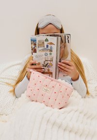 Rosa quilted veske med røde hjerte mønstre, hvit etikett med teksten "THE FLATLAY CO.", holdt av en person som leser et magasin mens de sitter på et mykt teppe.