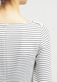 Randig topp i vitt och svart med fyrkantig halsringning och långa ärmar, med en synlig dragkedjestängning bak. Slät tygtextur.