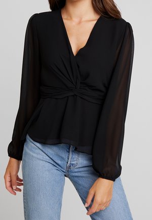 Frau trägt eine schwarze Bluse mit langen, transparenten Ärmeln und einem gedrehten Detail vorne, kombiniert mit hellblauen Jeans.