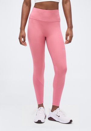 Nő, aki magas derekú rózsaszín leggingset és fehér Nike tornacipőt visel, áll egy egyszerű világos háttér előtt.