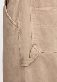 Beige tyg nära med detaljerad sömnad, ficka och ögla på ett plagg med en texturerad bomullsväv.