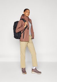 Chaqueta marrón resistente al agua, camiseta gris, pantalones beige, zapatos de senderismo marrones y una mochila negra, con un diseño ajustado y correas ajustables.