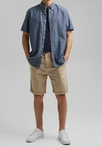 Koszula z krótkim rękawem w kolorze niebieskim z chambray, ciemna koszulka pod spodem, beżowe szorty z podwiniętymi nogawkami oraz białe sneakersy. Model stoi z rękami w kieszeniach.