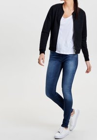 Kvinna klädd i en svart vadderad jacka, vit t-shirt, blå tighta jeans och vita sneakers, står med ett ben korsat framför det andra.