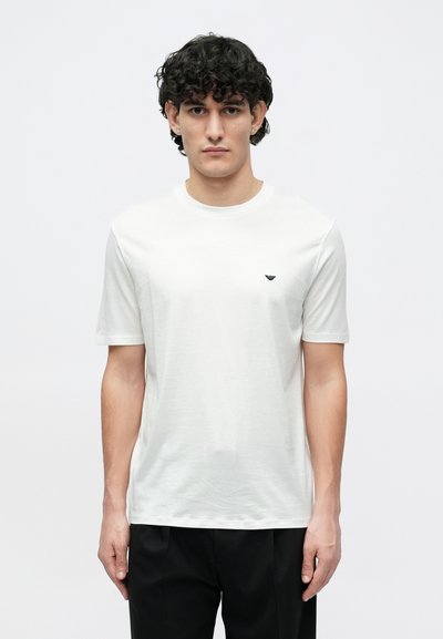 Fehér pamut póló rövid ujjakkal és kerek nyakkivágással. A bal mellrészen egy kis, sötét logó található. Simaságú anyag, normál szabás.