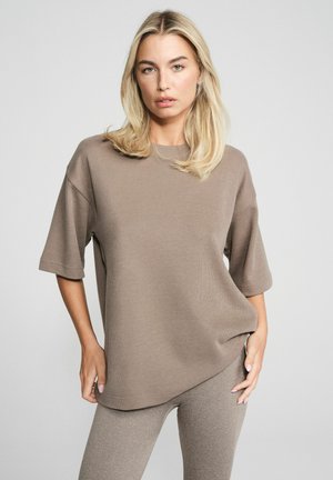 Blondynka w luźnej taupe'owej bluzce z krótkim rękawem i dopasowanych spodniach na jasnoszarym tle.