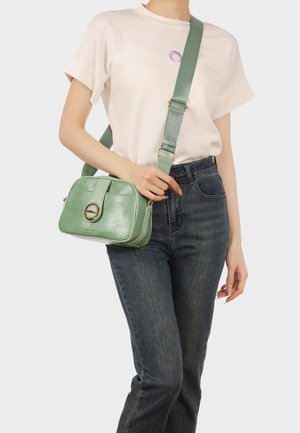 Persona che indossa una maglietta bianca e jeans scuri, porta una borsa a tracolla in pelle verde chiaro con una tracolla larga e una chiusura dorata.
