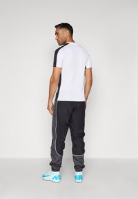 Männliches Sportoutfit mit einem kurzärmeligen Top in Weiß und Schwarz, schwarzen Hosen mit seitlichen Einsätzen und blauen Fußballschuhen.
