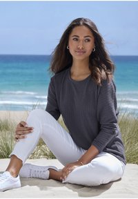 Junge Frau mit langen Haaren sitzt am sandigen Strand nahe Gras, trägt ein graues Langarmshirt, weiße Hosen und weiße Sneaker, mit dem Ozean im Hintergrund.