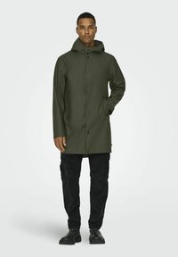 Cămașă impermeabilă de culoare verde măsliniu cu glugă, butoane frontale cu capse, buzunare laterale și croială dreaptă. Purtați peste pantaloni negri cargo și cizme.