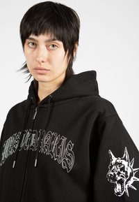 Μαύρη φούτερ φτιαγμένη από βαμβάκι με γραφικό μπροστά σε λευκό, που απεικονίζει διακοσμημένο κείμενο και ένα μεγάλο γραφικό κεφαλής σκύλου στο δεξί μανίκι.