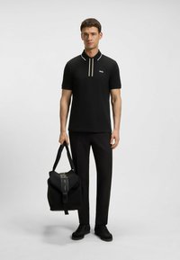 Schwarzes Poloshirt mit kontrastierendem weißen Kragen und Knopfleiste, kombiniert mit schwarzen Hosen. Das Model hält eine schwarze Reisetasche mit Reißverschluss.