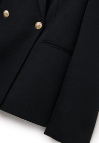 Blazer navy de tela texturizada, con botones dorados y un bolsillo en el pecho, corte ajustado con solapas afiladas y líneas limpias.