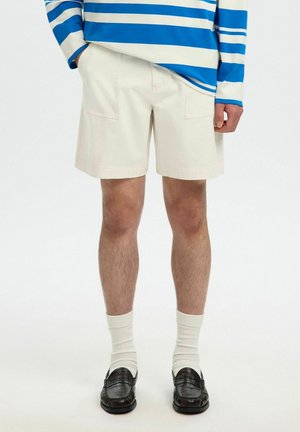 Mann trägt cremefarbene Shorts, weiße Socken, schwarze Loafers und ein blau-weiß gestreiftes Langarmhemd, die Hände entspannt an den Seiten.