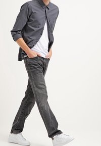 Camisa de botones gris sobre una camiseta blanca, acompañada de jeans negros desgastados y zapatillas blancas. Presenta un bolsillo en el pecho y mangas arremangadas.