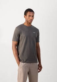 Mörkgrå bomulls t-shirt med korta ärmar, rundad halsringning och liten vit logga på bröstet, tillsammans med ljusbeige byxor.
